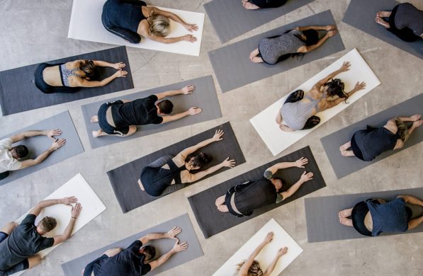 An aerial shot of a multi ethnic group of men and women practice yoga on on mats while wearing grey, black and white in an industrial setting. They are reaching forward in child's pose.